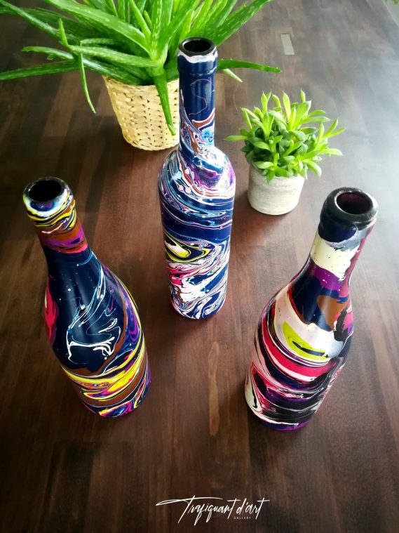 Trois bouteilles colorées décorées, entourées de plantes en pot sur une table en bois.