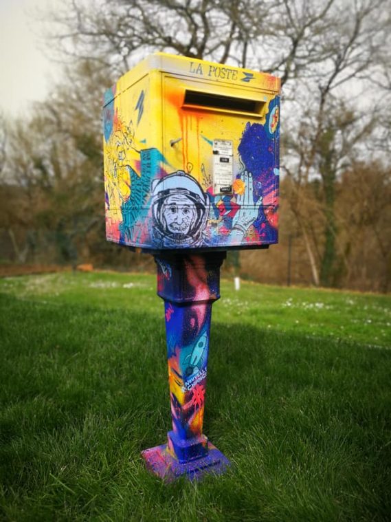 Borne aux lettres colorée avec des motifs graphiques et animaux, sur fond de verdure.
